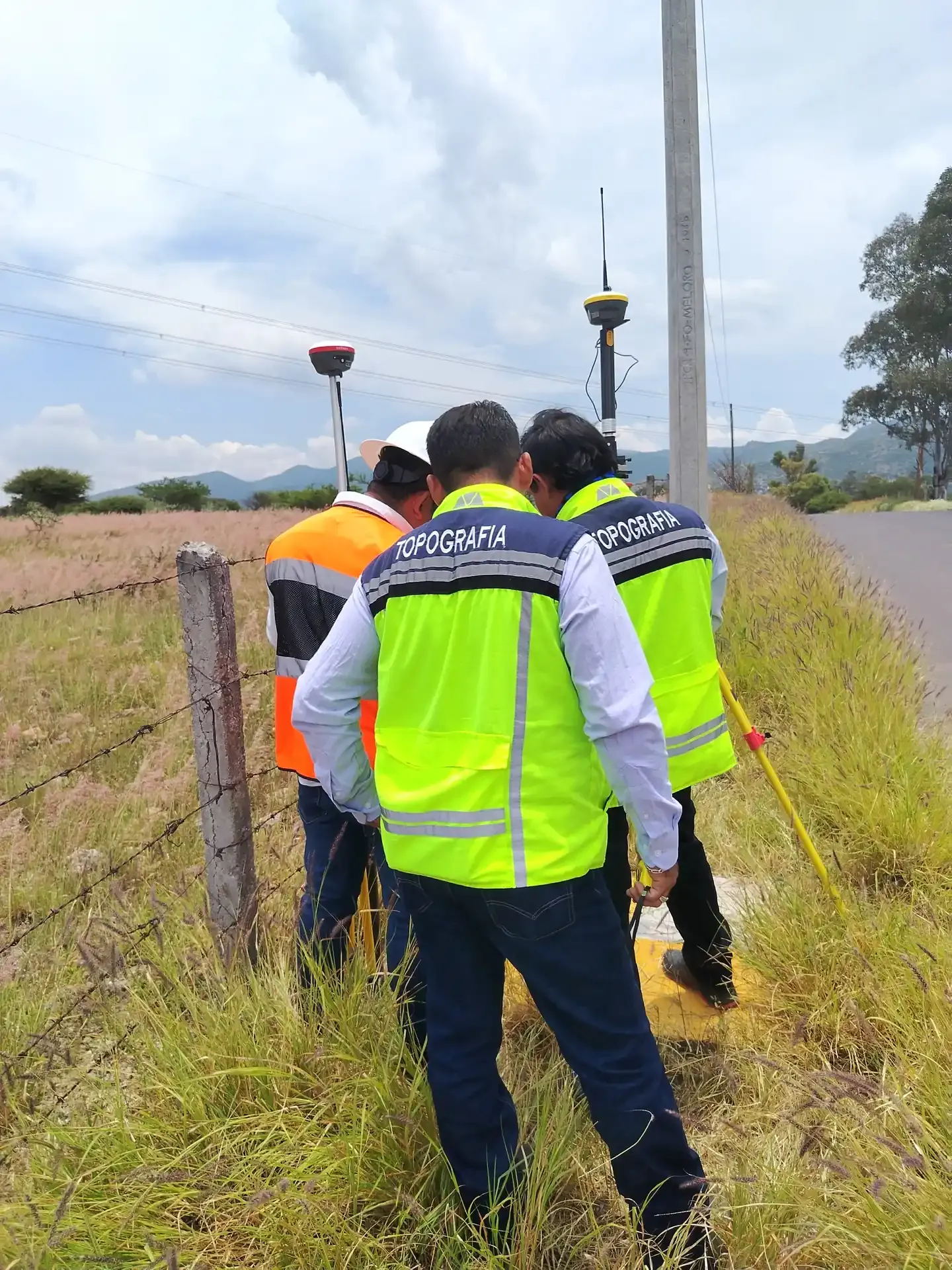 Brigada con receptores GNSS sobre bastón junto a una cerca, de espaldas
