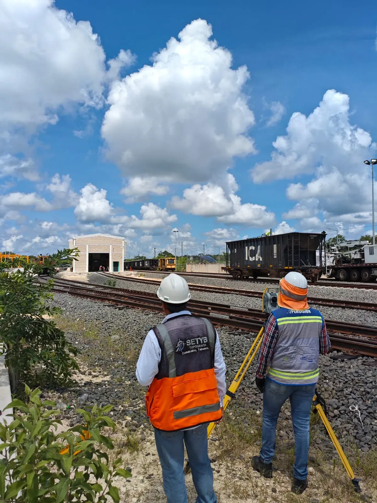 Dos topógrafos de espaldas con estación total frente a un patio ferroviario